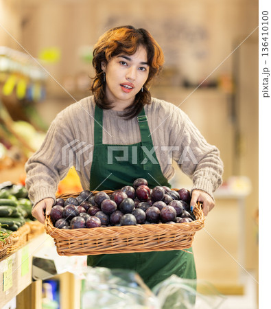 Young woman seller puts plums in store 136410100