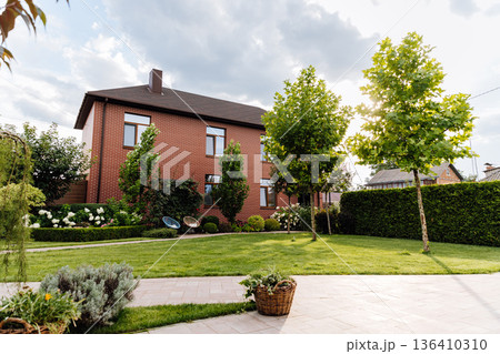 Brick family house with landscaped backyard, green lawn, trees and patio in sunlight Brick family house with landscaped backyard, green lawn, trees and patio in sunlight 136410310