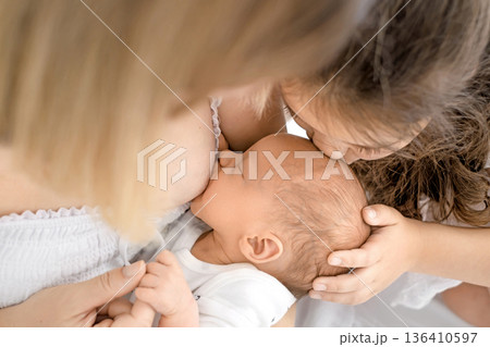 Tender moment of younger sister kissing newborn baby during breastfeeding. Family bonding, sibling love, baby health, care, support and early childhood development concept. Tender moment of younger sister kissing newborn baby during breastfeeding. Family bonding, sibling love, baby health, care, support and early childhood development concept. 136410597