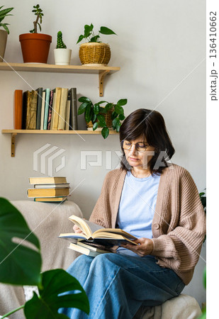 Woman in casual clothes relaxing while reading book at home. Plants and books fill shelves, creating peaceful atmosphere Woman in casual clothes relaxing while reading book at home. Plants and books fill shelves, creating peaceful atmosphere 136410662