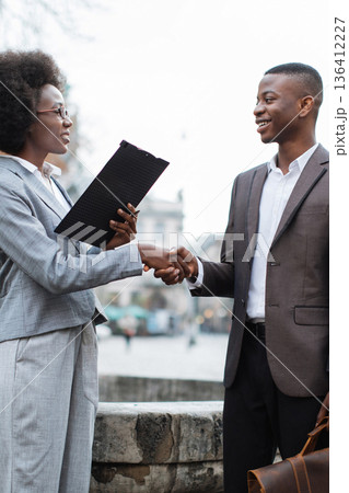 Two professionals, a man and a woman, shake hands outdoors while discussing business matters and reviewing documents 136412227