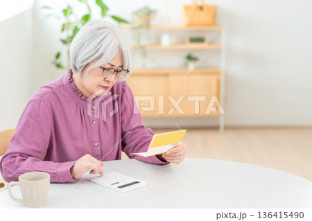 貯金通帳を見ながら電卓を使い計算するシニア女性（老後資金,生活費,年金） 136415490