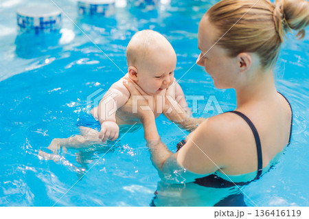 Mother bathing her baby in a swimming pool, holding and supporting the infant during water time. Tender bonding moment showing trust and care. Baby swimming, early development and healthy motherhood 136416119