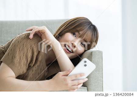 ソファに寝転がってスマホを操作する若い女性 ソファに寝転がってスマホを操作する若い女性 136420084