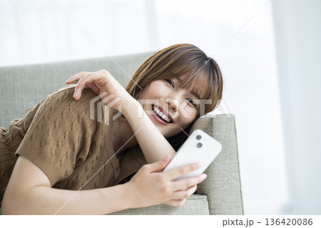 ソファに寝転がってスマホを操作する若い女性 ソファに寝転がってスマホを操作する若い女性 136420086