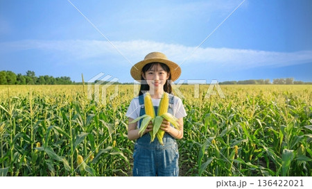 夏のトウモロコシ畑で収穫したトウモロコシを持って微笑む日本人少女 136422021