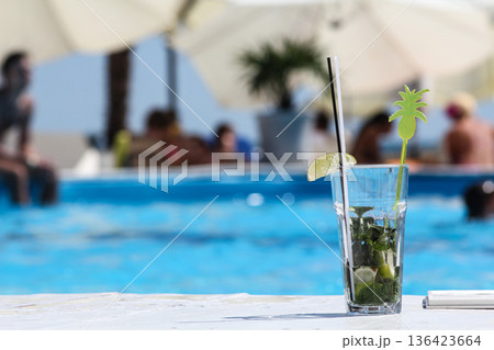Mojito cocktail against the backdrop of a blue pool on vacation, beach drinks, summer alcoholic cocktail mojito with ice, fresh mint and lime on table in cafe Mojito cocktail against the backdrop of a blue pool on vacation, beach drinks, summer alcoholic cocktail mojito with ice, fresh mint and lime on table in cafe 136423664