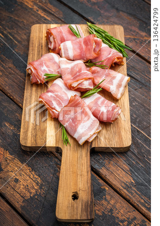 Rolled raw bacon pork rashers on rustic wooden board with rosemary and pepper ideal for cooking recipes and food styling content 136424799