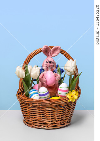 Multicolored Easter eggs in a basket with spring flowers: white tulips and yellow daffodils and pink Easter knitted bunny with pink egg on gray table. Easter concept Multicolored Easter eggs in a basket with spring flowers: white tulips and yellow daffodils and pink Easter knitted bunny with pink egg on gray table. Easter concept 136425220