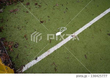 Charging cable lying on a green sports playground ground with a painted white line. Everyday tech object in a casual urban environment, themes of connectivity, technology, and modern lifestyle. Charging cable lying on a green sports playground ground with a painted white line. Everyday tech object in a casual urban environment, themes of connectivity, technology, and modern lifestyle. 136425777