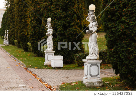 Marble statues on pedestals line a paved garden walkway with trimmed evergreen trees Marble statues on pedestals line a paved garden walkway with trimmed evergreen trees 136427052