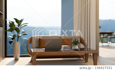 Laptop and books on glass top table and a wooden sofa chair aside window wall with mountain scenery. 136427331
