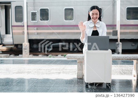 Smiling woman using laptop at train station, sitting on luggage, wearing headphones, waving during a video call. Smiling woman using laptop at train station, sitting on luggage, wearing headphones, waving during a video call. 136427390