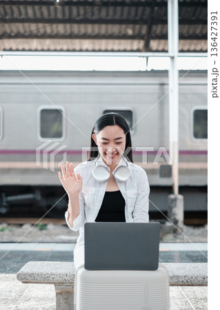 Young woman with headphones using a laptop at a train station, smiling and waving, representing modern travel and communication. Young woman with headphones using a laptop at a train station, smiling and waving, representing modern travel and communication. 136427391