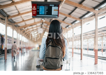 A young woman with a backpack stands on a train station platform, looking at the digital display board. 136427393