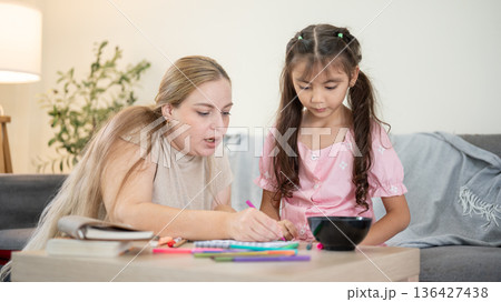Caucasian mother holding color pen drawing with kid daughter at table aside sofa in living room. Caucasian mother holding color pen drawing with kid daughter at table aside sofa in living room. 136427438