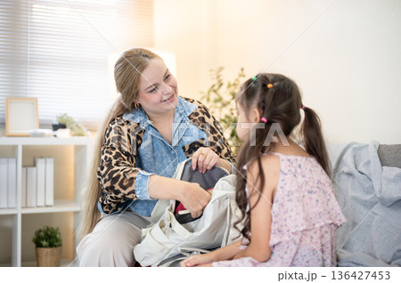 Caucasian mother organizing school bag for asian kid daughter while sitting on sofa in a living room Caucasian mother organizing school bag for asian kid daughter while sitting on sofa in a living room 136427453