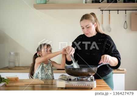 Kid daughter holding arm of white mother while cooking in pan on stove standing at kitchen counter. 136427479
