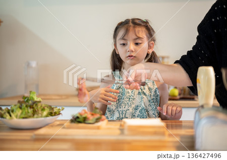Asian daughter looking at mother putting bacon over bread making sandwich at kitchen cooking counter 136427496
