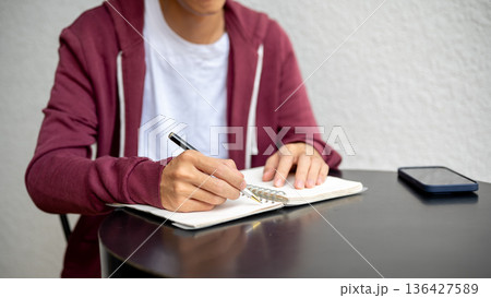 A man or student holding pen writing in notebook aside smartphone on black table in cafe or library. 136427589