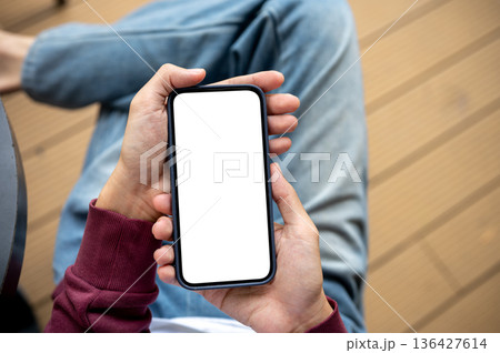 A man holding blank screen smartphone while sitting cross legged at table in cafe or coffee shop. 136427614