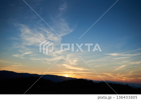 Sun sets behind rolling hills, casting warm, golden glow across sky. Wispy clouds, tinged with hues of orange and yellow, stretch across deepening blue sky. Serene and expansive landscape. Sun sets behind rolling hills, casting warm, golden glow across sky. Wispy clouds, tinged with hues of orange and yellow, stretch across deepening blue sky. Serene and expansive landscape. 136433805