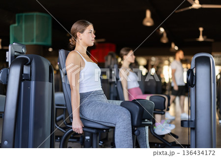 Slender athletic girl work out muscles of legs and thighs on simulator in fitness club Slender athletic girl work out muscles of legs and thighs on simulator in fitness club 136434172