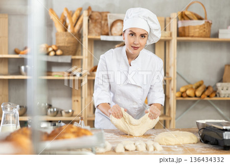 Skilled smiling young girl professional baker in white uniform standing at work table and kneading dough during working day in bakery 136434332