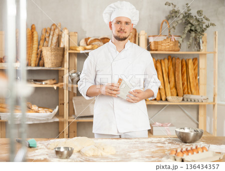 Male baker stands near work table in bakery, resting while working. Male baker stands near work table in bakery, resting while working. 136434357