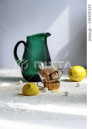 Emerald green glass jug and yellow lemon on white table against green wall.  136435325