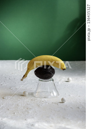 Surreal balance composition with yellow banana on top of avocado and glass stand.  136435327