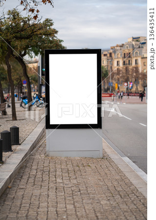 Blank vertical street billboard mockup showcasing a clean white space, ideal for advertising and marketing campaigns, positioned on a city sidewalk next to a road with blurred urban background 136435411