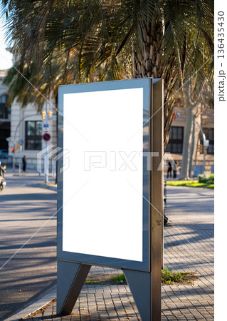 Blank outdoor advertising billboard mockup on a city street, providing copy space for promotion and marketing campaigns under warm sunlight next to a palm tree 136435430