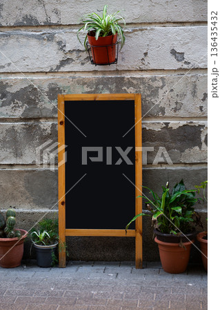 Blank chalkboard stand with wooden frame leaning on a rustic concrete wall, surrounded by potted plants, creating an inviting street cafe or restaurant outdoor display mockup Blank chalkboard stand with wooden frame leaning on a rustic concrete wall, surrounded by potted plants, creating an inviting street cafe or restaurant outdoor display mockup 136435432
