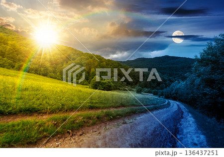 country road down the hill through the field. day and night time change concept. lovely countryside scenery of mountainous area on summer solstice with sun and moon. remote pathway in rural highland 136437251