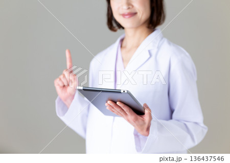 タブレットを持って人差し指を立てる白衣姿のミドル女性看護師 タブレットを持って人差し指を立てる白衣姿のミドル女性看護師 136437546
