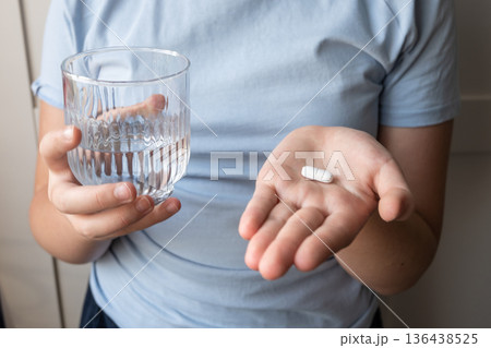 Pill and glass with water in the hand of child. Healthcare from pharmacy. Kid sit at home because of illness. Pill and glass with water in the hand of child. Healthcare from pharmacy. Kid sit at home because of illness. 136438525
