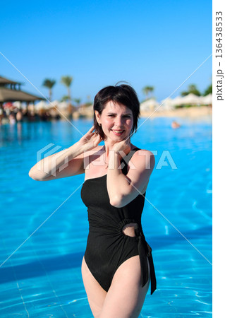 A striking woman in a black bikini stands knee-deep in a pool 136438533