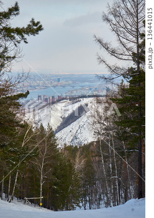 Winter landscape with snow-covered mountains, forest, river, bridge, cityscape on a gloomy day. Scenic view for travel, skiing, nature. Krasnoyarsk, Siberia 136441015