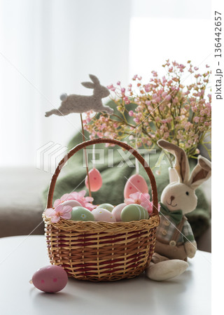 Easter Basket with Ester Eggs, Plush Bunny and Spring Decor on White Table intoCozy Living Room Easter Basket with Ester Eggs, Plush Bunny and Spring Decor on White Table intoCozy Living Room 136442657