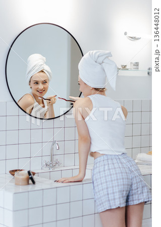 Blonde with a white towel on her head brushes her teeth in front of a mirror in a white bathroom. 136448012