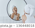 Blonde with a white towel on her head brushes her teeth in front of a mirror in a white bathroom. 136448018