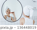 Blonde woman with a white towel on her head brushes her teeth in front of a round mirror in a white bathroom. 136448019