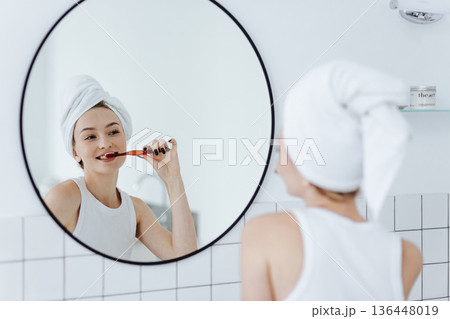 Blonde woman with a white towel on her head brushes her teeth in front of a round mirror in a white bathroom. Blonde woman with a white towel on her head brushes her teeth in front of a round mirror in a white bathroom. 136448019