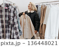 Young Smiling Blonde Woman Trying on shirt, standing between the Rails with clothes 136448021