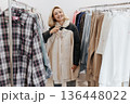 Young Smiling Blonde Woman Trying on shirt, standing between the Rails with clothes 136448022