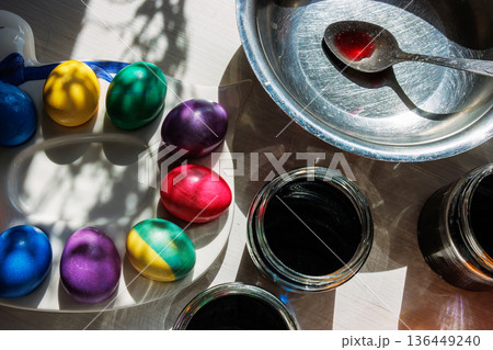 Closeup of brightly dyed eggs on a palette beside glass jars of dark dye and a metal bowl with a stained spoon on a sunlit tabletop. 136449240