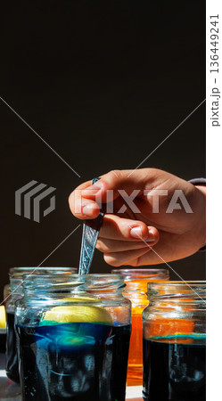 Vertical close up of a hand holding a spoon with an egg submerged in a blue food dye jar  on dark background with copy space 136449241