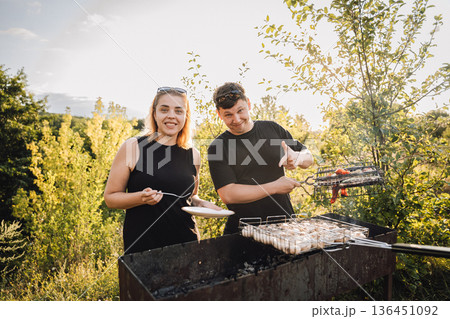 Couple grilling vegetables outdoors at sunset, tasting food at summer barbecue Couple grilling vegetables outdoors at sunset, tasting food at summer barbecue 136451092