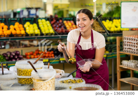 In vegetable department,female seller in apron put pickled olives in disposable container with ladle In vegetable department,female seller in apron put pickled olives in disposable container with ladle 136453800